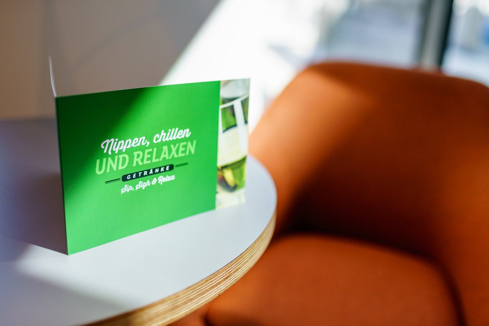 Grüne Getränkekarte mit Aufschrift 'Nippen, chillen UND RELAXEN' steht auf einem weißen Tisch neben einem orangefarbenen Sessel